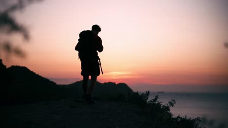 silhouette shot of a young backpacker standing in the cliff of a mountain at sunset and watching the sea