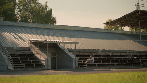 young woman sitting alone on stadium bleachers under metal roof structure, immersed in thought, surrounded by greenery and architecture, with trees and mast in background