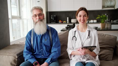 retrato de una mujer feliz en un abrigo blanco con un estetoscopio y con una tableta en sus manos que está sentada en un sofá junto con un hombre mayor con una barba gris exuberante en una camisa azul en un apartamento moderno durante un examen en casa