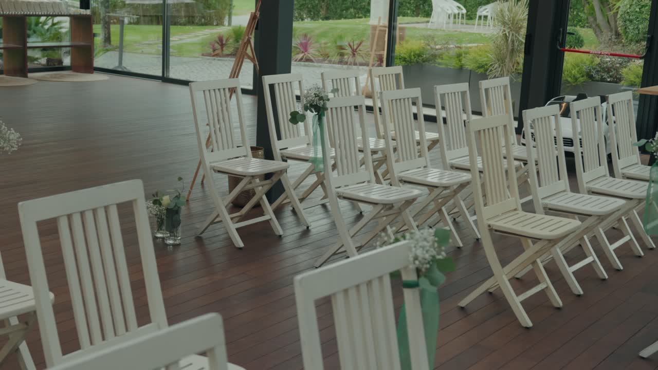 Chaises pliantes en bois blanc alignées en rangées sur un parquet avec de petites décorations florales dans des vases en verre.