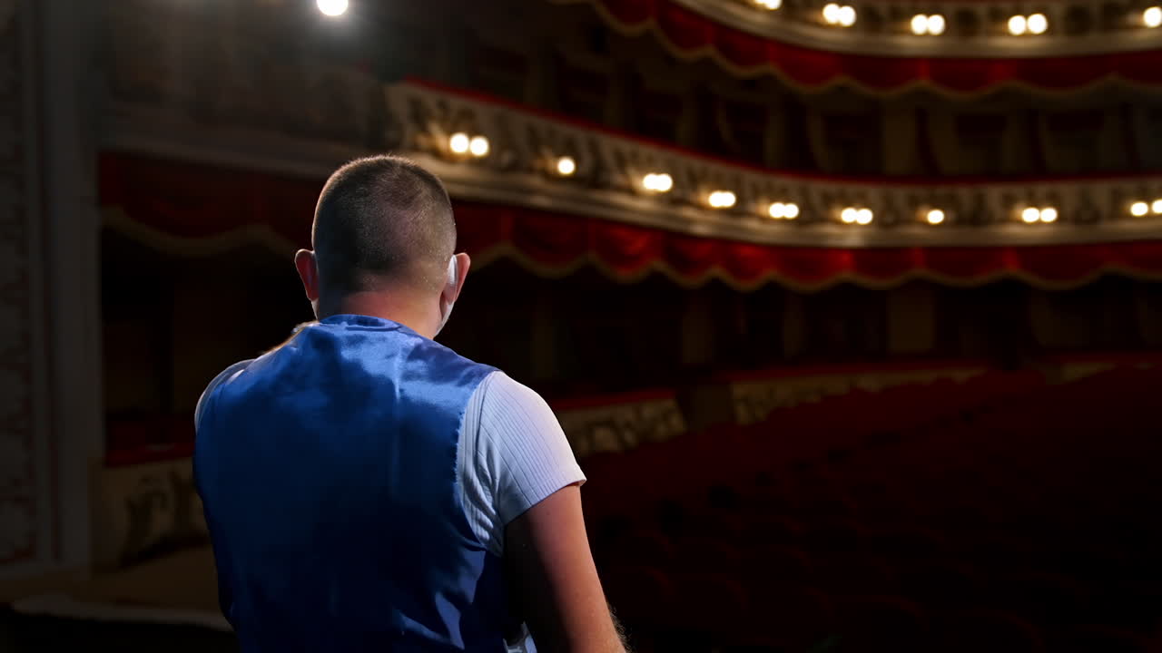 Showman entertains the audience under the light, front view. Big stage. Serious performer speaks from the stage in protective mask. Hands in pockets.