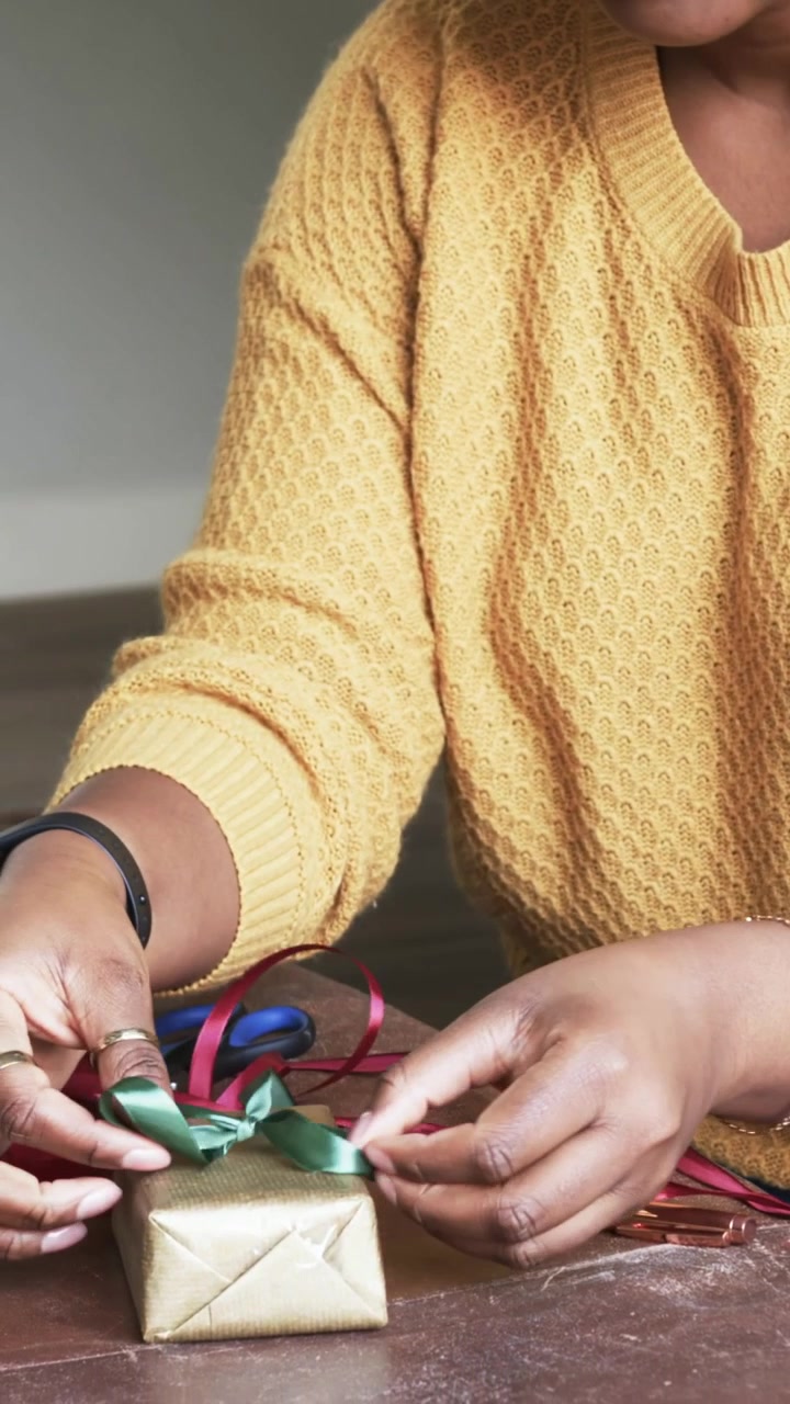 Video verticale: Donna con un maglione color senape che sceglie un nastro verde e forma un fiocco a casa, decorando una scatola dorata.