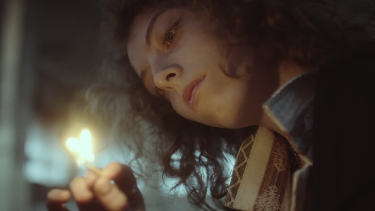 A young woman gazes at a lit matchstick in a dim setting