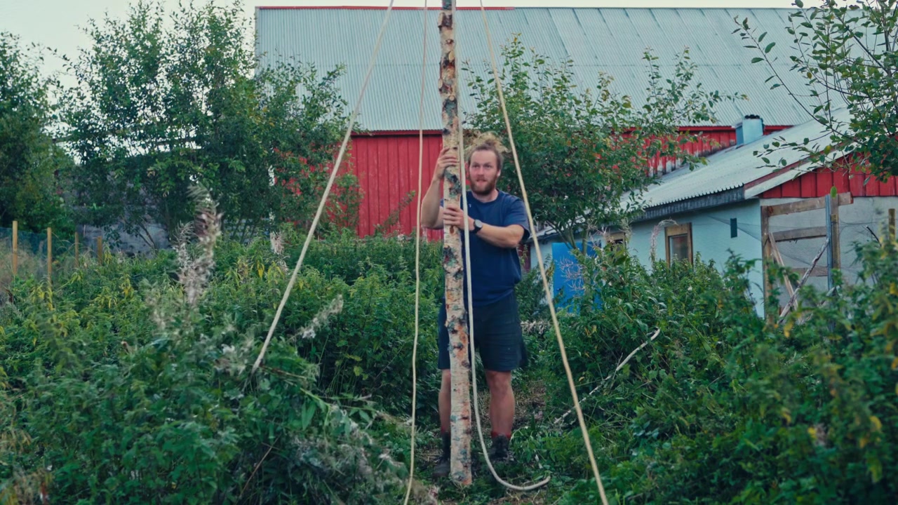 Mann som holder opp bjørkestang med tau i landlig hage. Monterer DIY tipi-ramme. Vidvinkelbilde