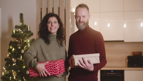 front view of a couple smiling at camera and holding christmas present in a room decorated with a christmas tree