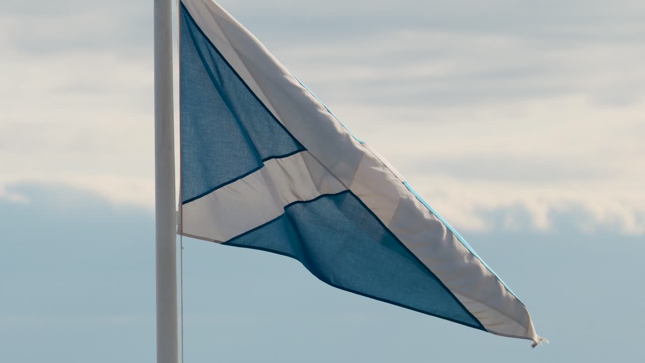 Blue and white saltire flag flutters dynamically on flagpole, daylight, steady camera, outdoor setting