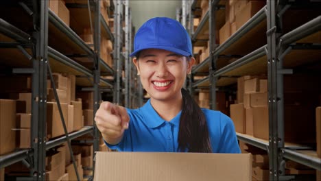 primer plano de una mensajera asiática en uniforme azul sonriendo y haciendo un gesto honesto mientras entrega una caja en el almacén