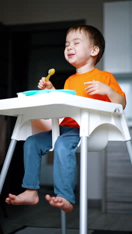 cheerful caucasian toddler sits at high chair chewing his food. low angle view on the kid enjoying his meals. vertical video.