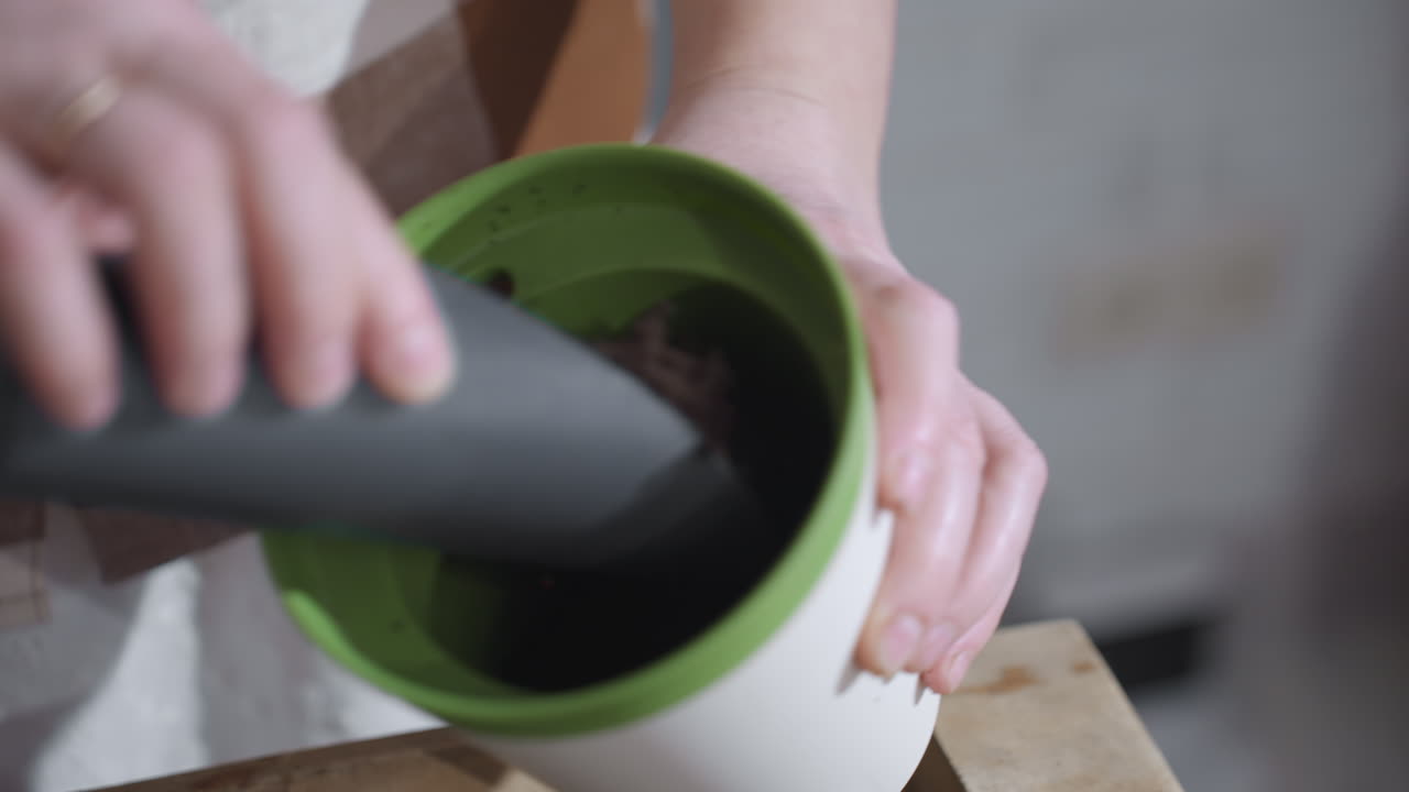 Lady putting potting soil from wooden bowl into plastic planter on glass table under soft indoor light showcasing mindful gardening ritual with textured earth details and focused nurturing hands