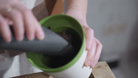 lady putting potting soil from wooden bowl into plastic planter on glass table under soft indoor light showcasing mindful gardening ritual with textured earth details and focused nurturing hands