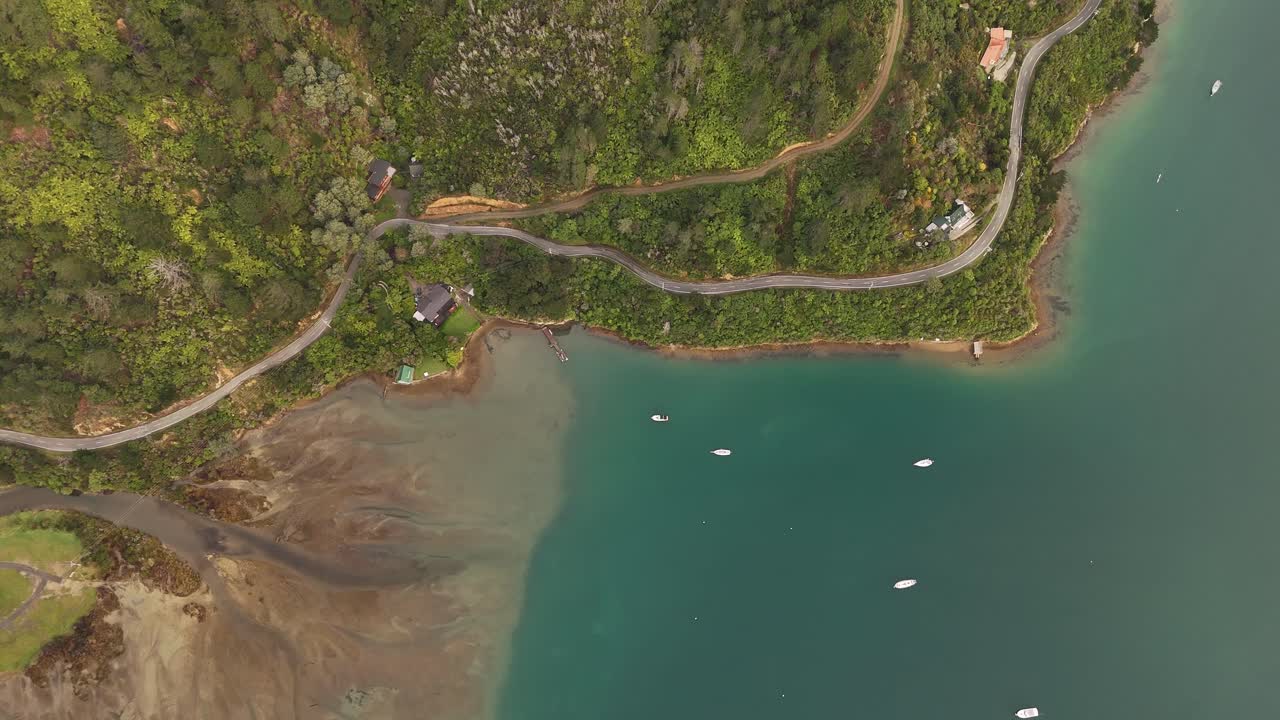 Coastal road through lush green forest, bay with anchored boats and houses along shore, Picton, New Zealand. Aerial top-down orbiting