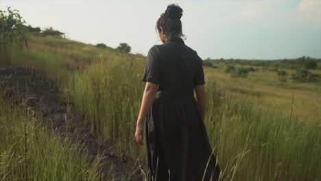 A-young-black-haired-woman-in-a-long-black-dress-walks-along-a-grassy-path