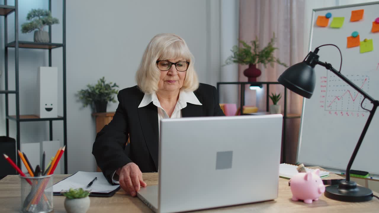 Smiling mature business woman sits at workplace office desk opening laptop computer start working