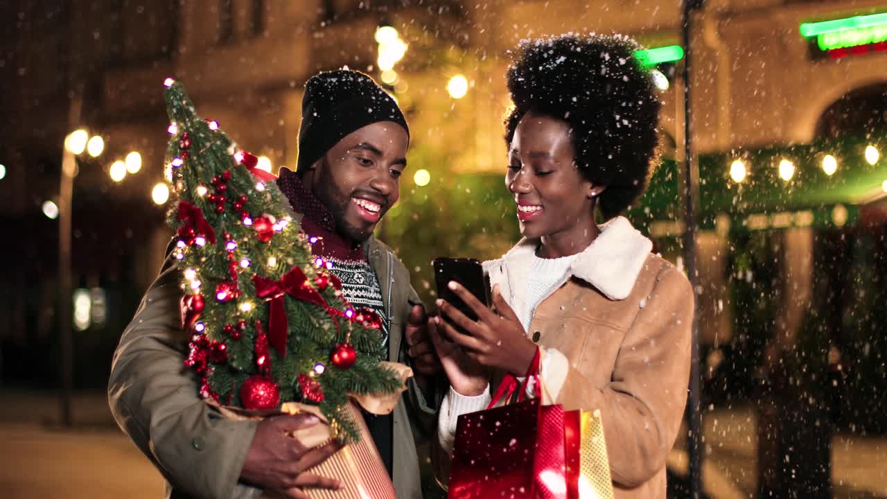 vista de cerca de una alegre pareja afroamericana hablando y viendo algo por teléfono mientras nieva en la calle en navidad