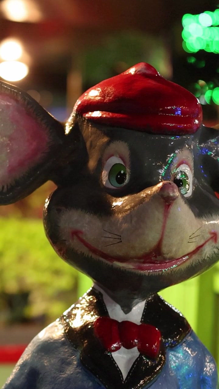 A vertical close-up shot of a smiling mouse character statue under colorful nighttime lights.