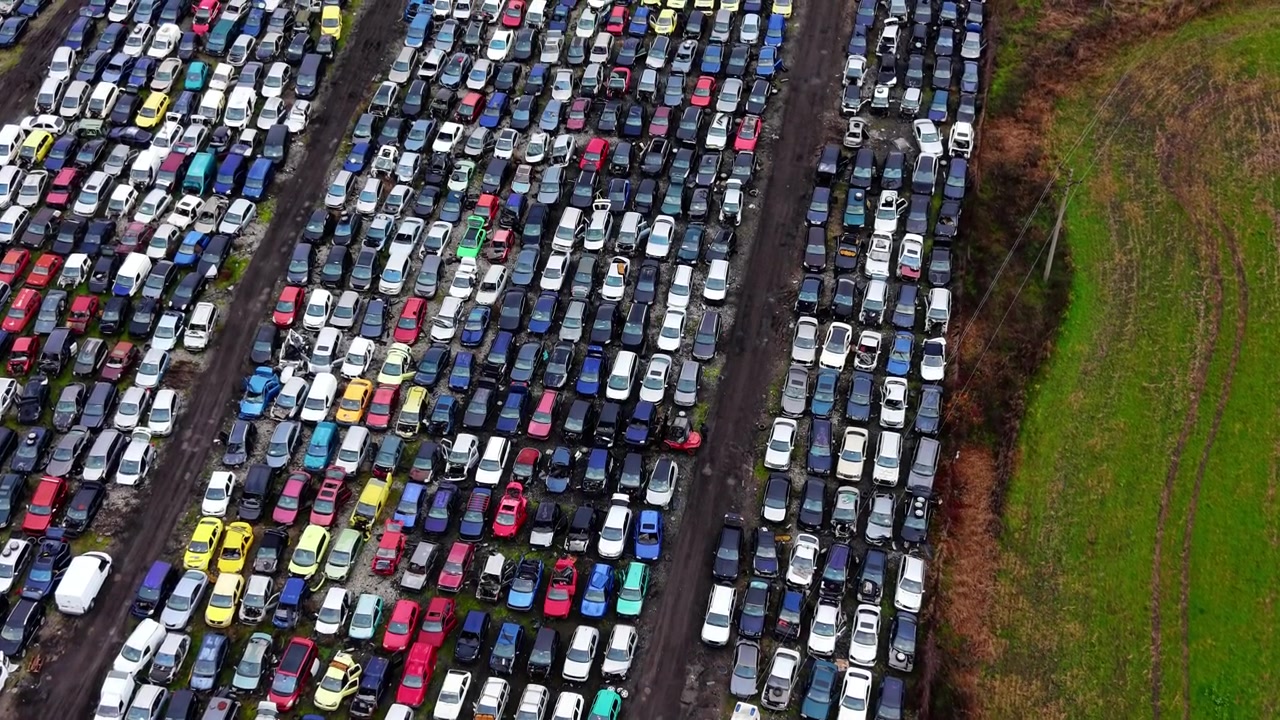 Filas de vehículos siniestrados en un desguace al aire libre junto a tierras de cultivo, Bulgaria. Vista de dron elevada y angulada de coches multicolores en líneas densas junto a un campo verde y líneas eléctricas.