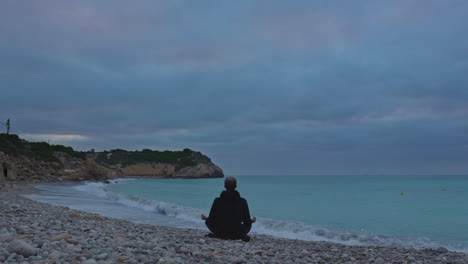 Time-lapse-captures-man-sitting-on-stoney-beach-meditating-by-the-seaside-in-Sitges,-Spain