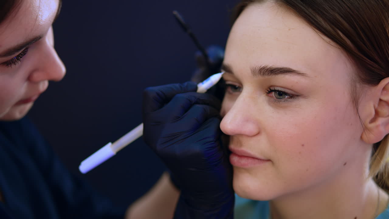 Beautiful young lady is in beauty salon. Brow master drawing the lines near the eyebrows. Close up.