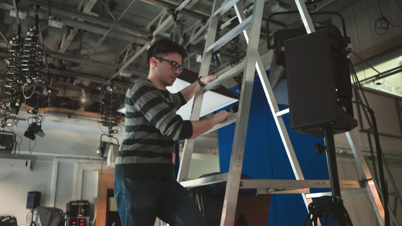 Studiotechniker klettert Leiter hoch, um Beleuchtungs- und Kameraausrüstung im Broadcast-Studio anzupassen, bereitet sich auf Dreharbeiten in Produktionsumgebung vor, justiert Ausrüstung für optimales Setup