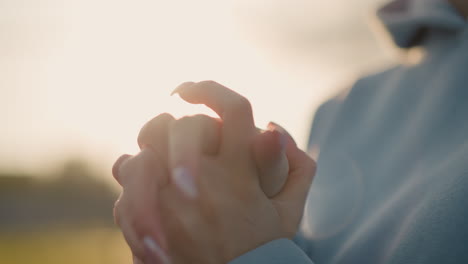 close-up of woman with polished nails performing wrist twist in blue sweater, sunlight creating warm glow around hands, highlighting movement and elegance in serene outdoor setting