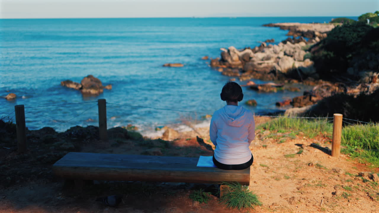 Woman with headphones on her head sitting on a wooden bench with a view of the sea