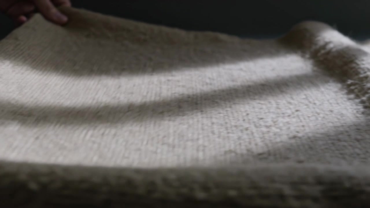 Dita che entrano dall'alto a sinistra e tirano, levigando una maglia beige su un tavolo grigio, appiattendo con fasce di luce.