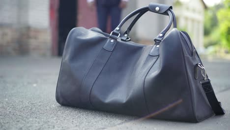 close-up of black leather travel bag outdoors with blurred african american man walking at background and taking suitcase. unrecognizable businessman in formal suit on business trip in urban slum.