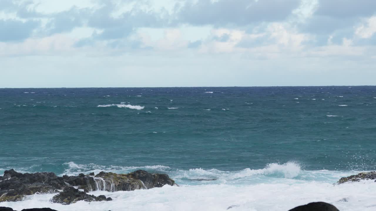 Wide ocean scene with foamy waves breaking against rugged rocks, sea spray rising dramatically into the air under a cloudy horizon