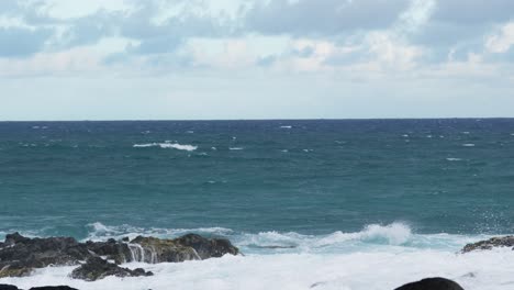 wide ocean scene with foamy waves breaking against rugged rocks, sea spray rising dramatically into the air under a cloudy horizon