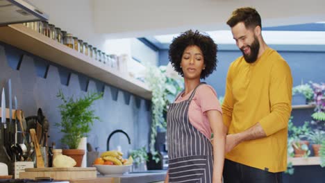 video de una feliz pareja diversa poniendo un delantal y hablando en la cocina en casa, con espacio para copiar