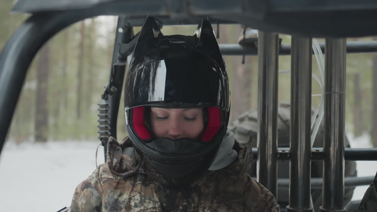 Un pilota in ATV UTV che sorride sotto il casco, con una giacca mimetica, guance arrossate, espressione rilassata dopo una corsa adrenalinica, incorniciato da pini innevati, luce soffusa, con un'atmosfera di sollievo post-avventura e cameratismo.