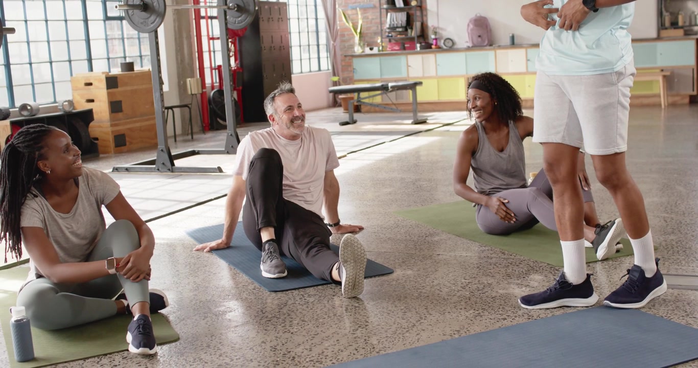 Diverse class turning toward trainer stepping in, giving guidance, smiling, shifting on mats in gym
