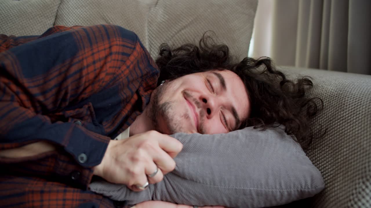 A happy brunette guy with curly hair in a checkered shirt lies down on a soft pillow and with a smile on his face rests at home on the sofa in a modern apartment
