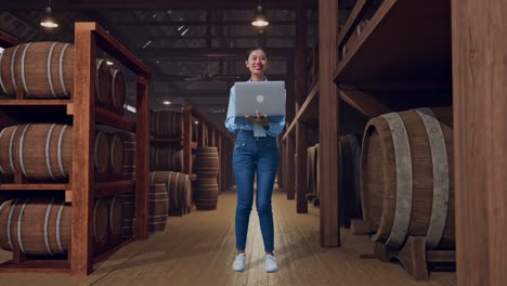 full body of an asian female worker standing with her laptop in cellar of winery, she observes by looking around before she come to concentrating with her laptop