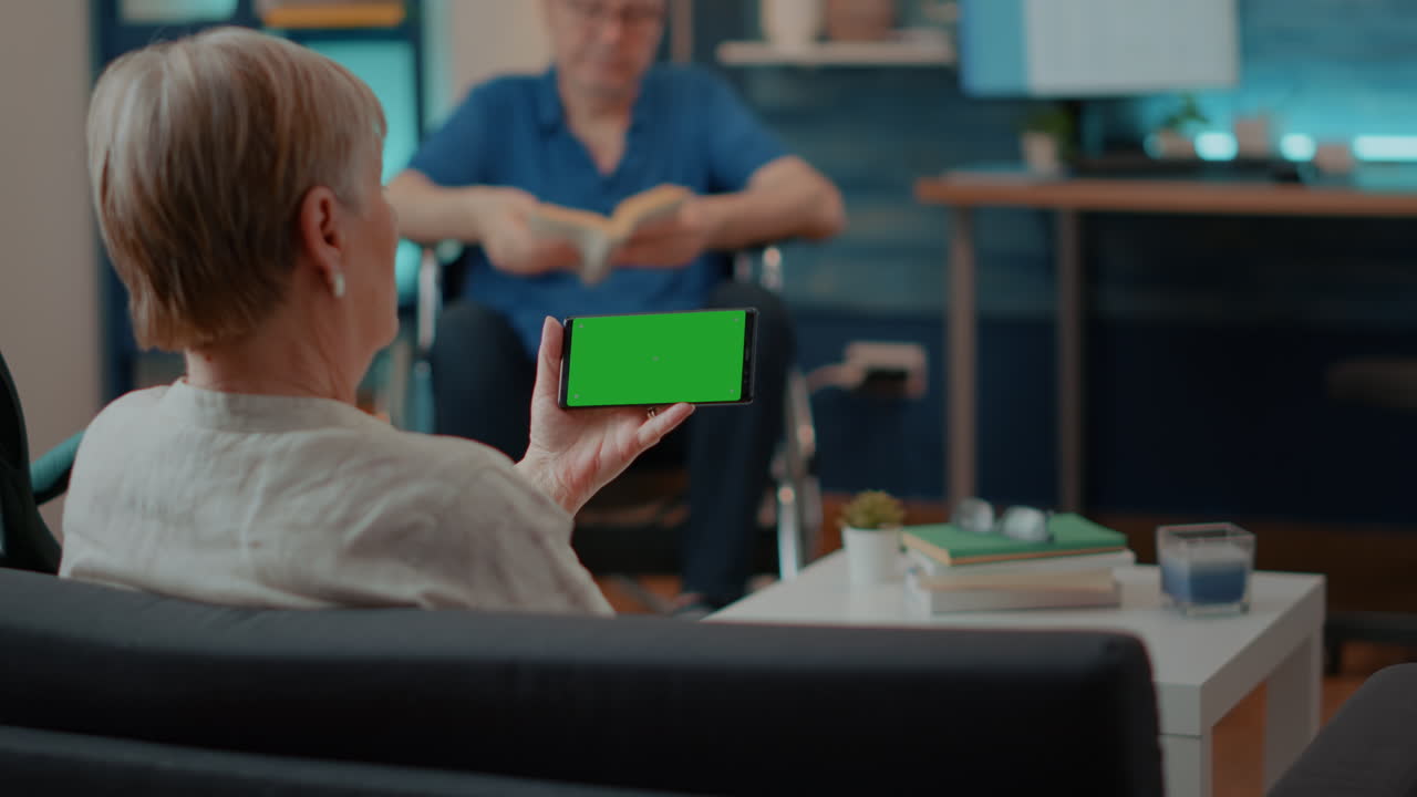 Senior Woman Using Smartphone with Green Screen While Husband Reads Book
