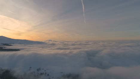 Antena,-Toma-De-Drones,-Sobre-Nubes,-Frente-Al-Bosque-Nevado,-Picos-De-Montaña-Y-Colores-De-Puesta-De-Sol