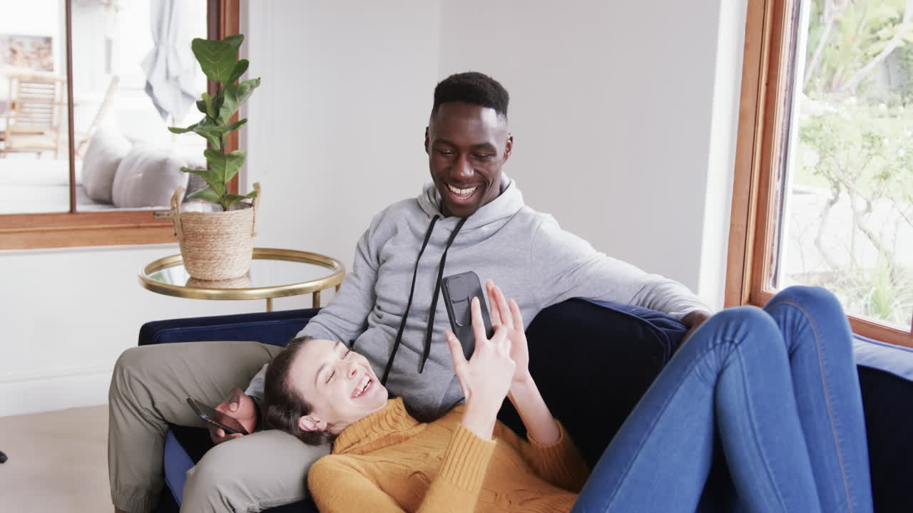 Happy diverse couple sitting and lying on sofa and using smartphones in home,copy space