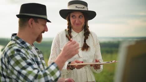 a sketcher wearing a black hat and checkered shirt hands over a palette to a woman in a white dress beside him, standing together in a peaceful grass field