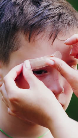Sad boy with injury on his eyebrow. Woman's hands putting small white adhesive plaster on a wound outdoors. Close-up. Vertical video