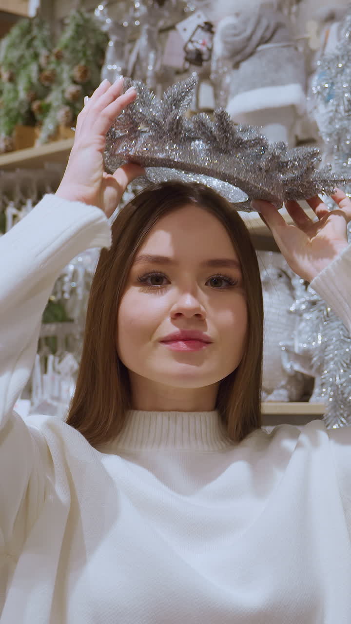 Lady picks sparkling Christmas decoration from shelf in festive decor store and playfully places it on her head, enjoying festive shopping in beautiful holiday atmosphere