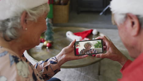 pareja caucásica mayor usando un teléfono inteligente para una videollamada de navidad con un amigo sonriente en la pantalla