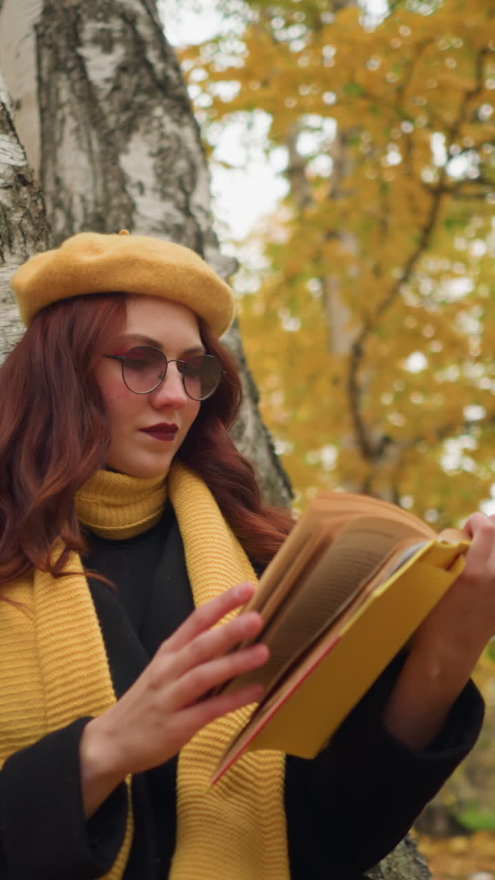 joven contemplativa pasa por su libro, sentada sola en el parque, a punto de abrir otra página, con boina amarilla y silenciador, el fondo presenta árboles con hojas doradas de otoño