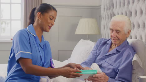 female care worker in uniform helping senior man at home in bed with medication