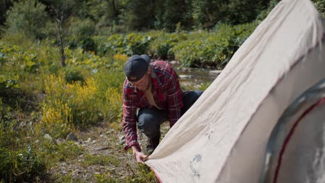 an overview of a tourist in glasses with a beard who, in special clothing for hiking in red, puts up a tent in a forest area near the river