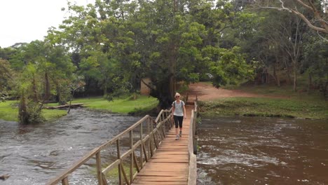 aerial tracking shot of a young blonde woman walking across a wooden footbridge over a river