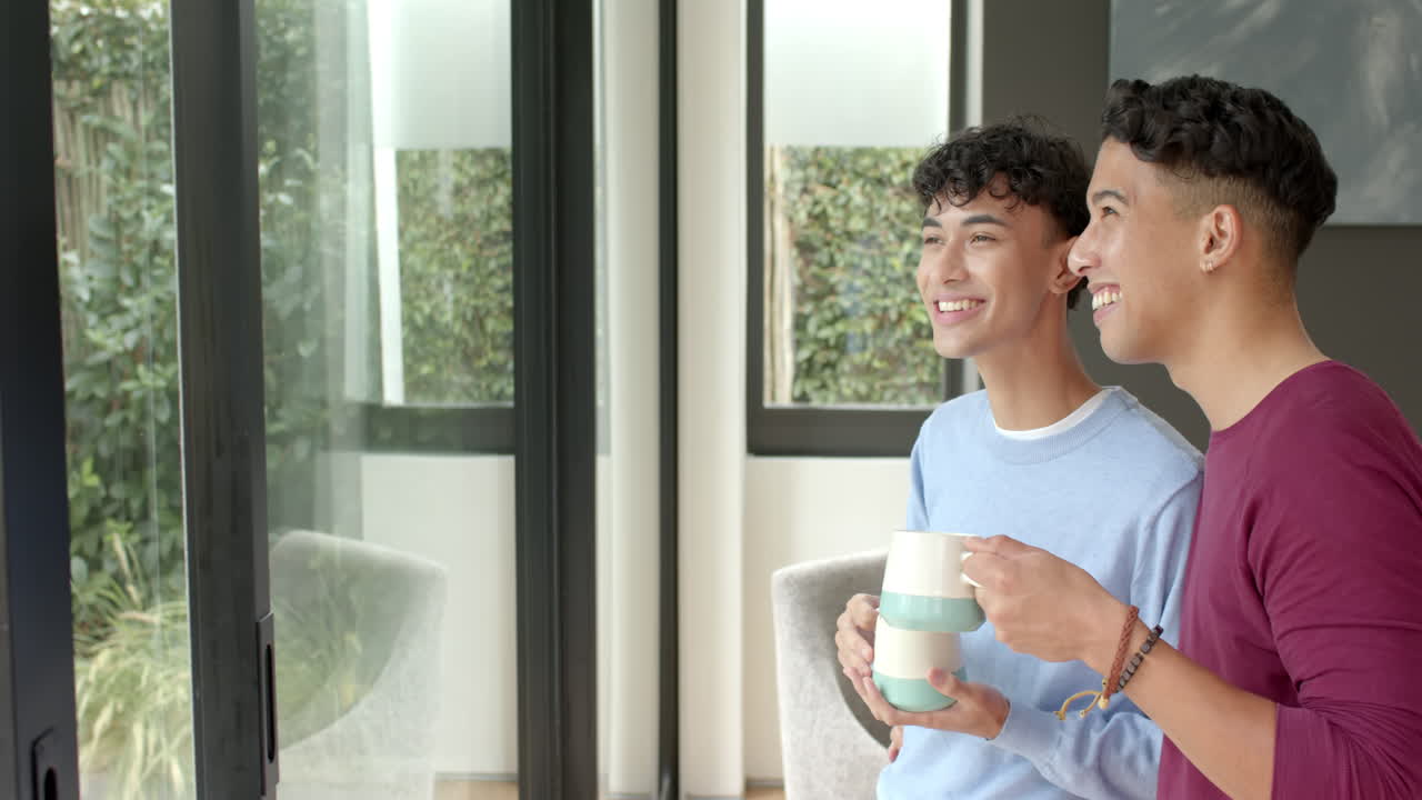 Smiling multiracial gay couple enjoying coffee by window at home
