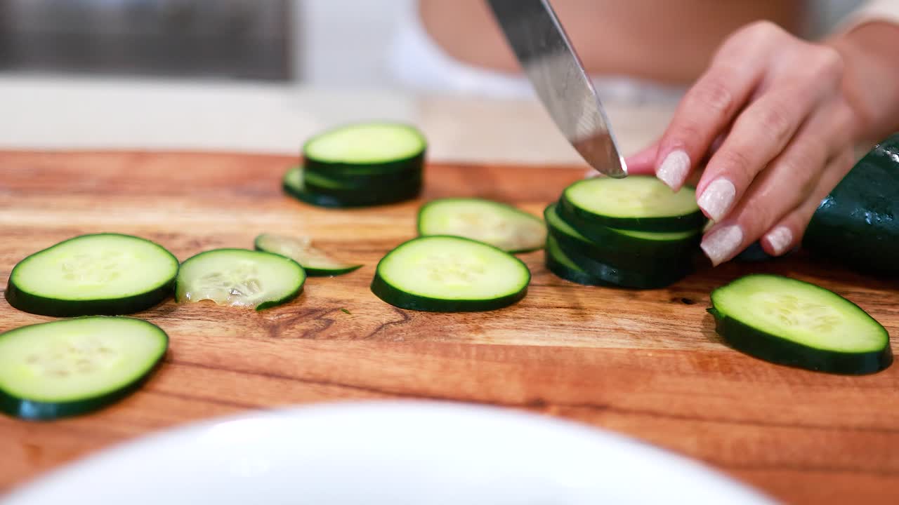 Hands slicing cucumber on a wooden board in a bright kitchen. Close-up, natural lighting, focus on fresh produce