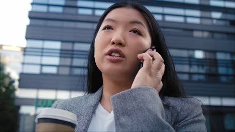 Business-Chinese-woman-having-a-call-and-holding-a-cup-of-coffee-while-standing-on-the-street