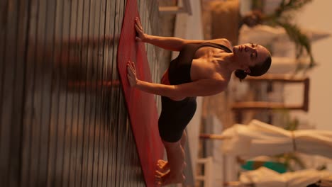 vertical video: a brunette girl with tied hair in a black sports uniform does yoga and gets into a cat pose on a sunny beach covered with boards during golden sunrise in summer
