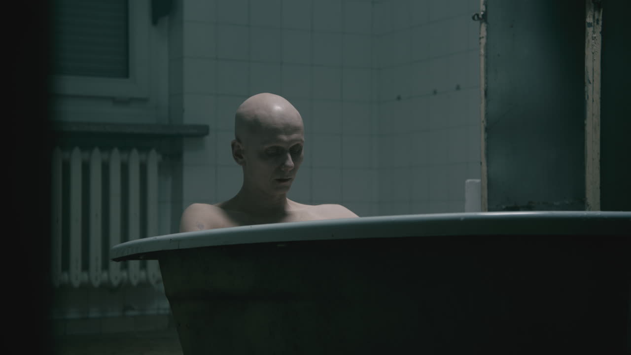 Pale bald skinny man takes a bath in the creepy old bathroom at night